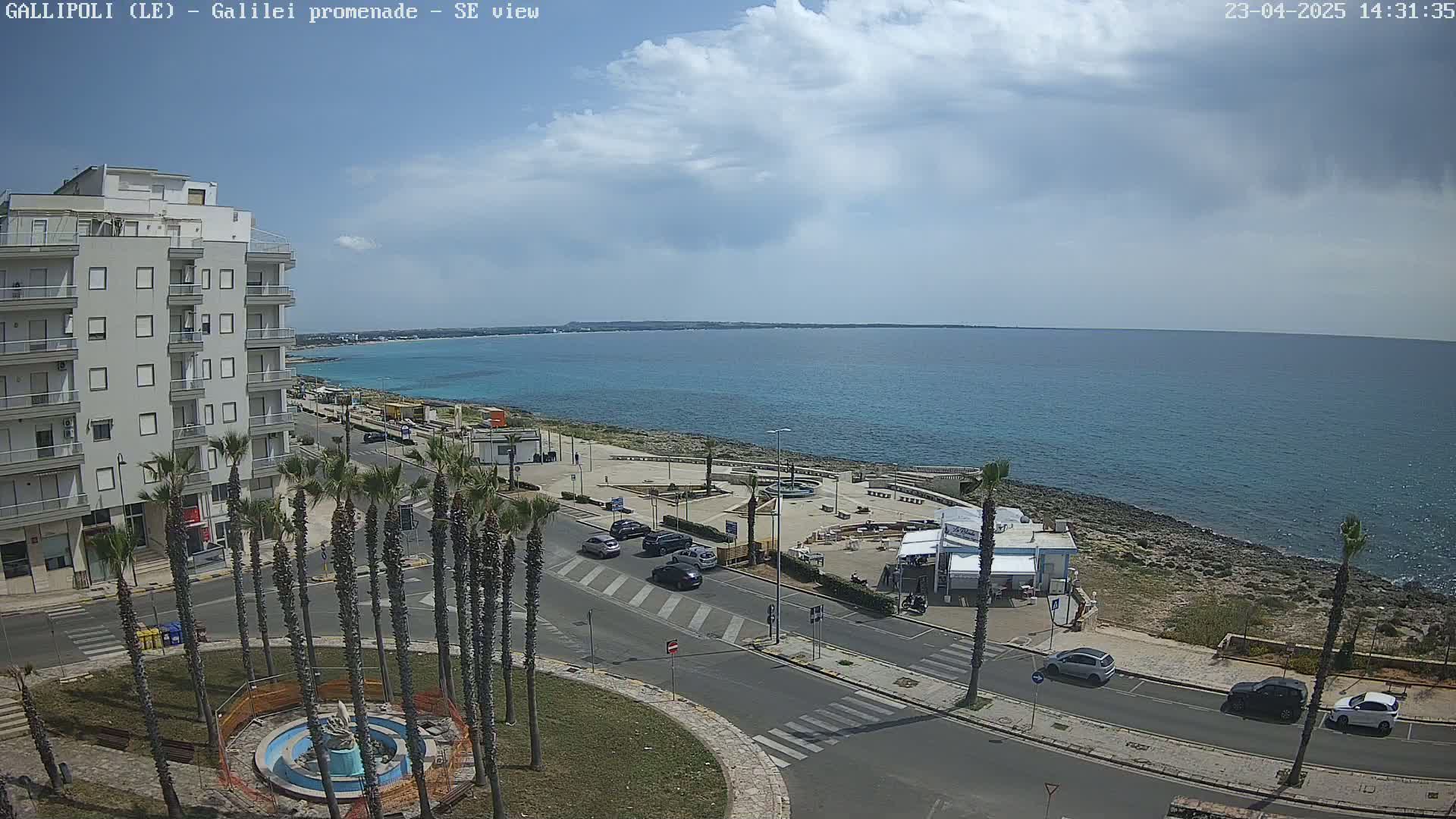 A partly cloudy day reveals a seaside town with a coastal road, palm trees, a building, and a calm blue ocean.