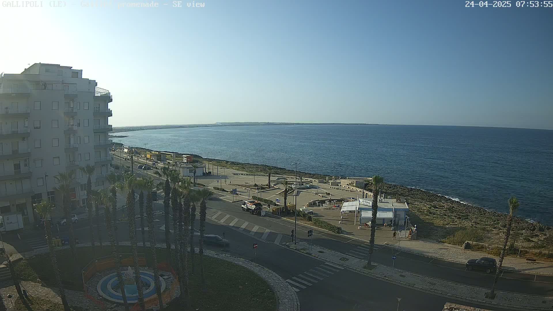 An elevated view shows a seaside town on a clear, sunny day, with a calm ocean extending to the horizon, a roadway curving around a circular plaza with palm trees, and a multi-story building.