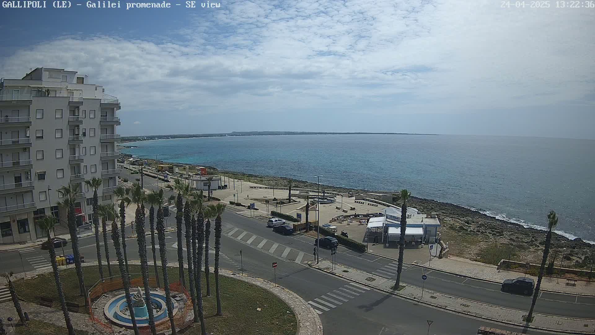A partly cloudy day overlooking a coastal town with a promenade, palm trees, a fountain, a few cars, and a building next to a calm ocean.