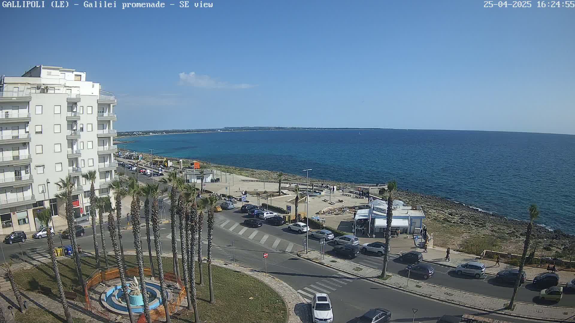 A seaside town on a sunny day shows a calm blue ocean, a paved coastal promenade with some people and beachside kiosks, and palm trees lining a street next to a multi-story building.