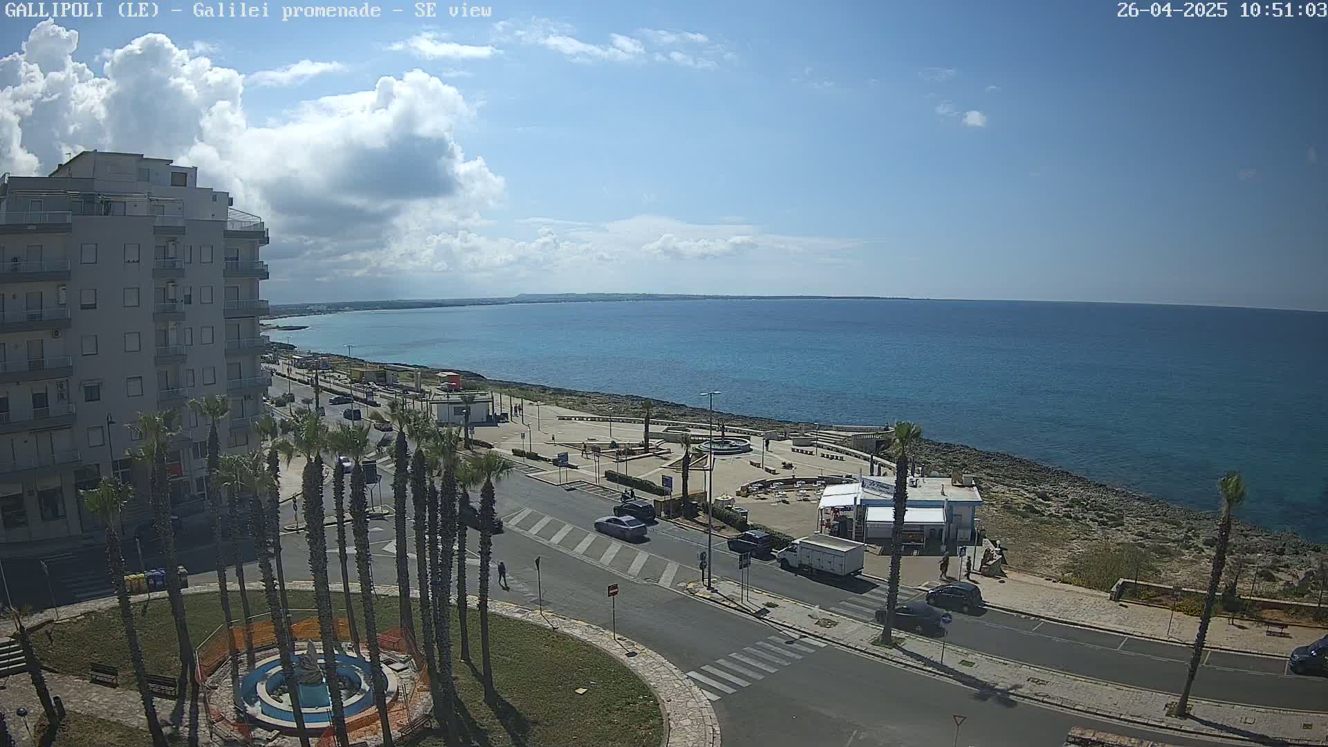 A partly cloudy sunny day overlooking a seaside town with a promenade, palm trees, buildings, cars, and a fountain.