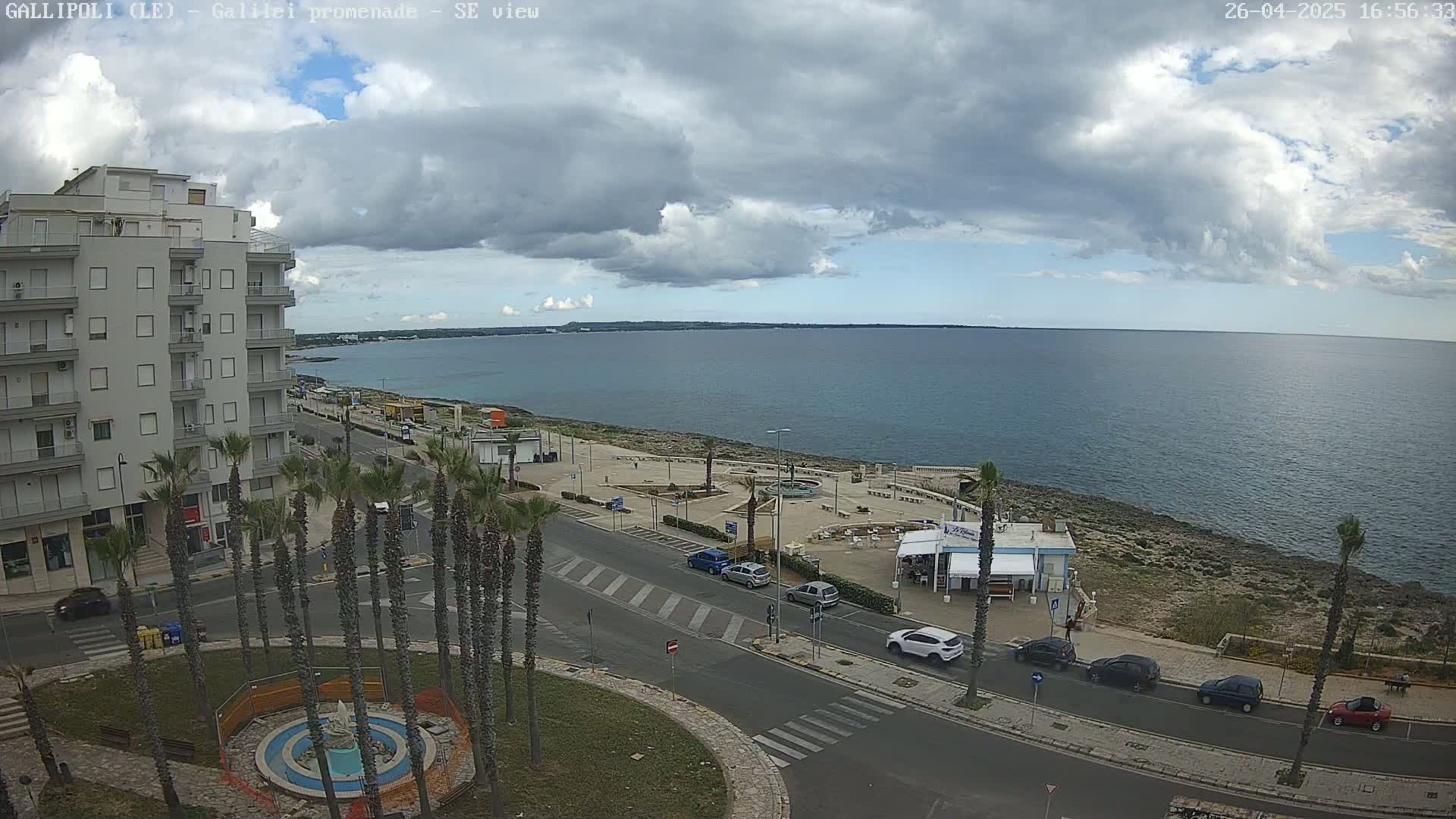 A partly cloudy day shows a coastal city scene with palm trees lining a street next to a calm ocean, where cars are parked along the road near a waterfront restaurant.