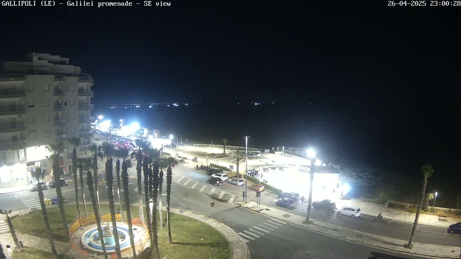 A nighttime aerial view shows a coastal town with cars parked along a street near a body of water under a clear, dark sky.