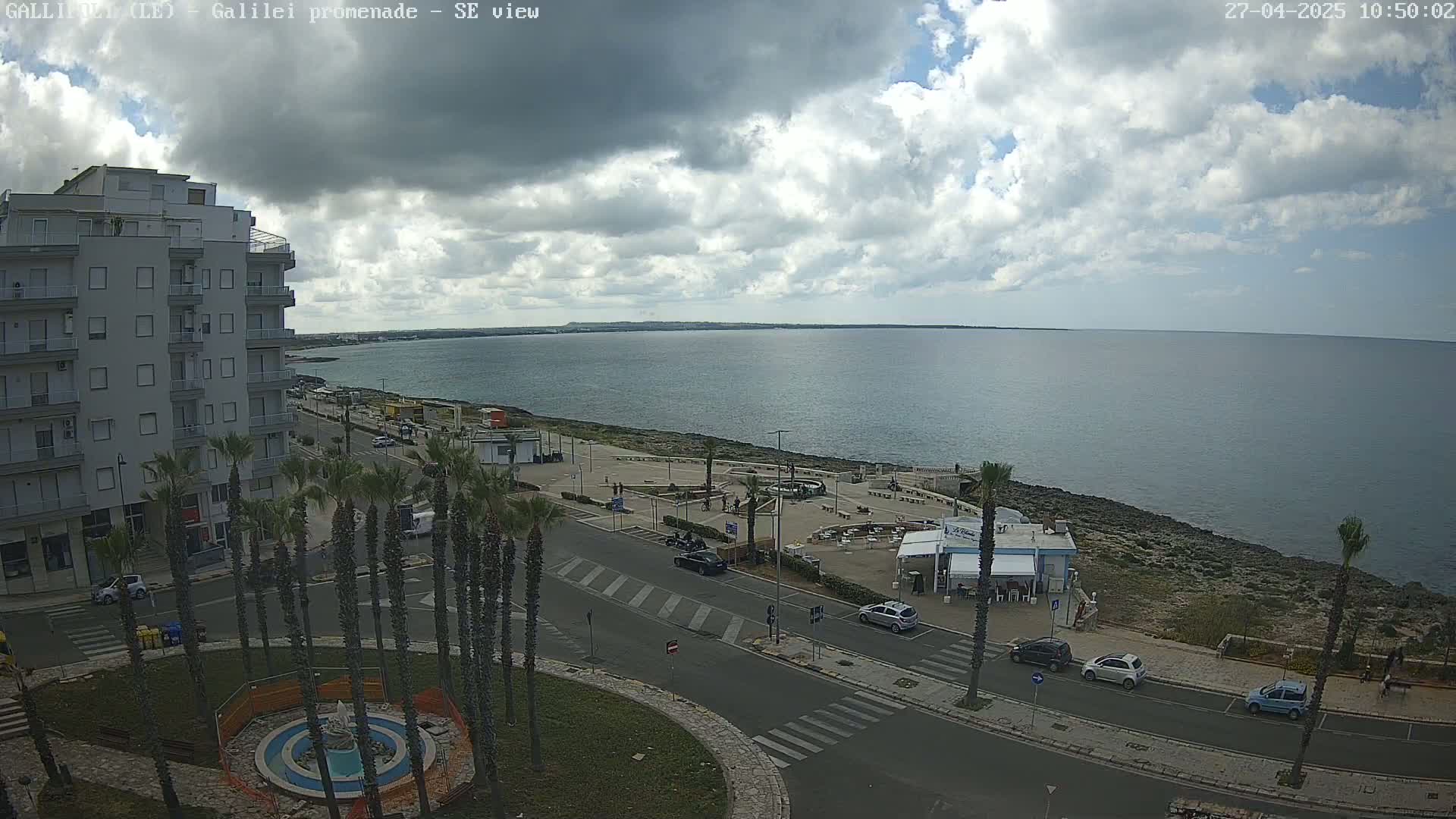 A partly cloudy day overlooks a seaside promenade with palm trees, buildings, parked cars, and a small plaza near the ocean.