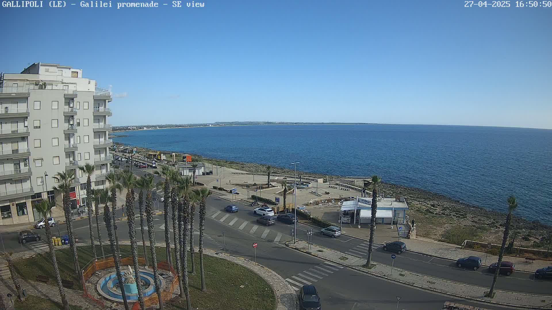 A clear, sunny day overlooks a coastal city scene with a calm ocean, palm trees lining a roadway, and several cars parked along the street.