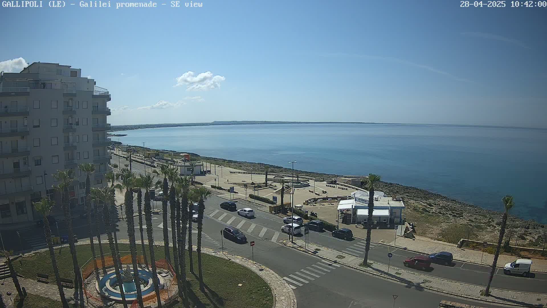 A seaside town scene on a sunny day shows a multi-story building, palm trees, a roadway with cars, and a promenade overlooking a calm blue sea.