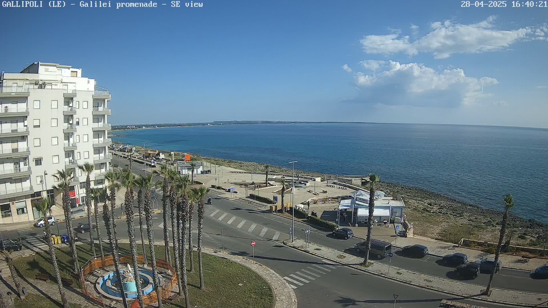 A partly cloudy day overlooks a seaside town with palm trees lining a road next to a calm ocean.