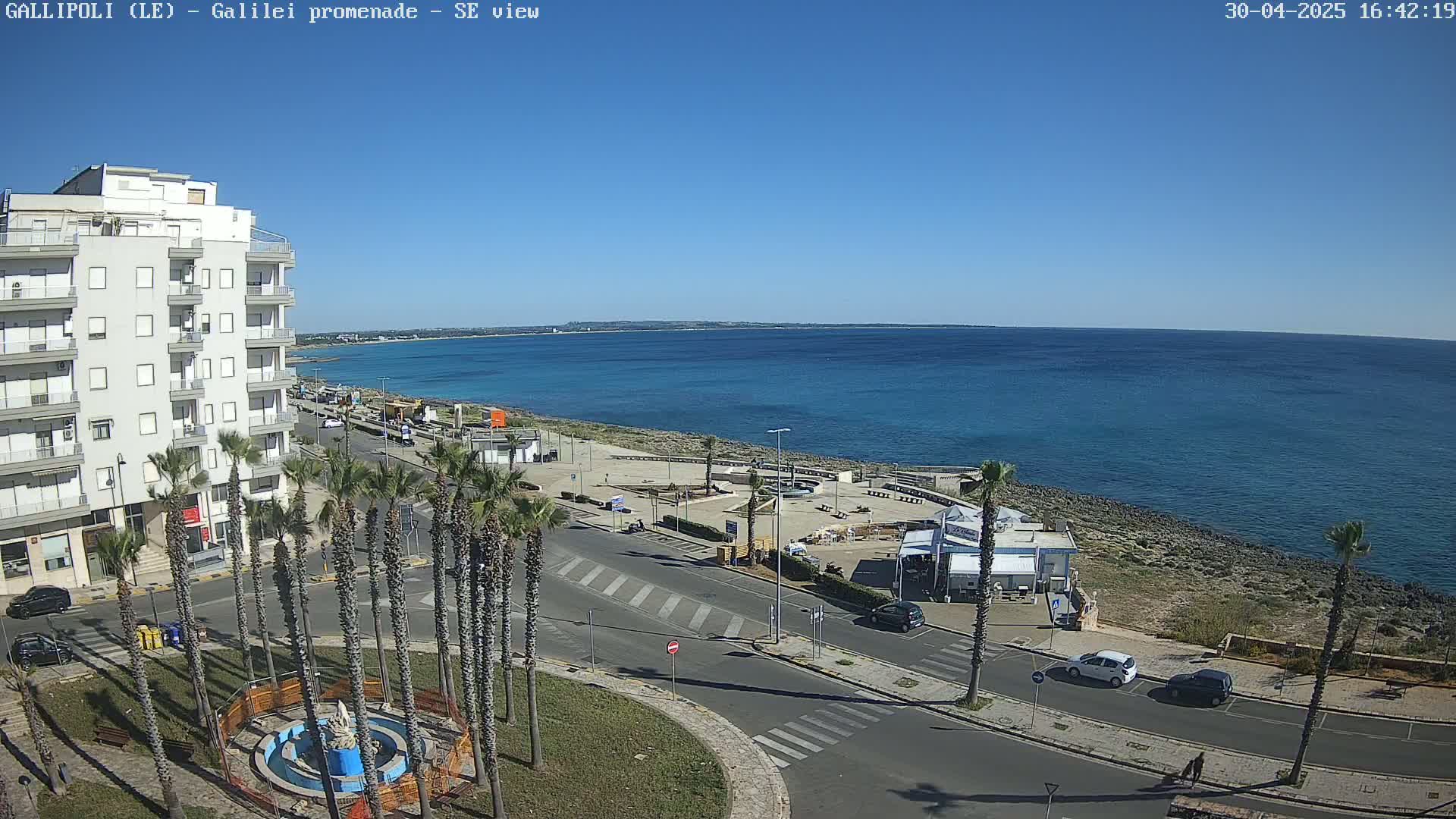 A seaside town on a clear, sunny day, with palm trees lining a paved road that leads to a calm blue ocean.