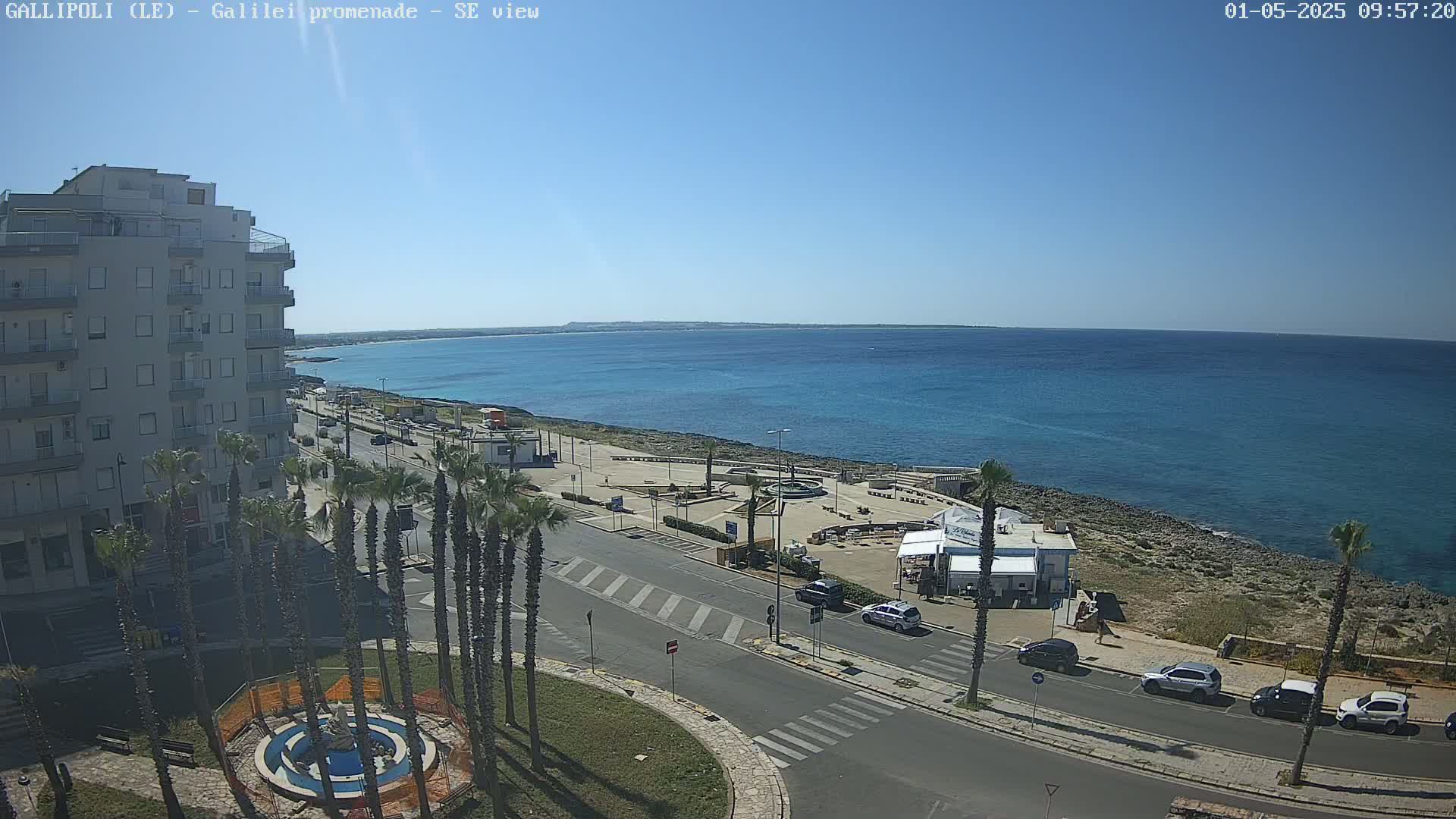 A seaside town scene on a clear, sunny day shows a multi-story building, palm trees lining a road leading to a waterfront promenade with a small restaurant, and a calm blue sea extending to a distant shoreline.