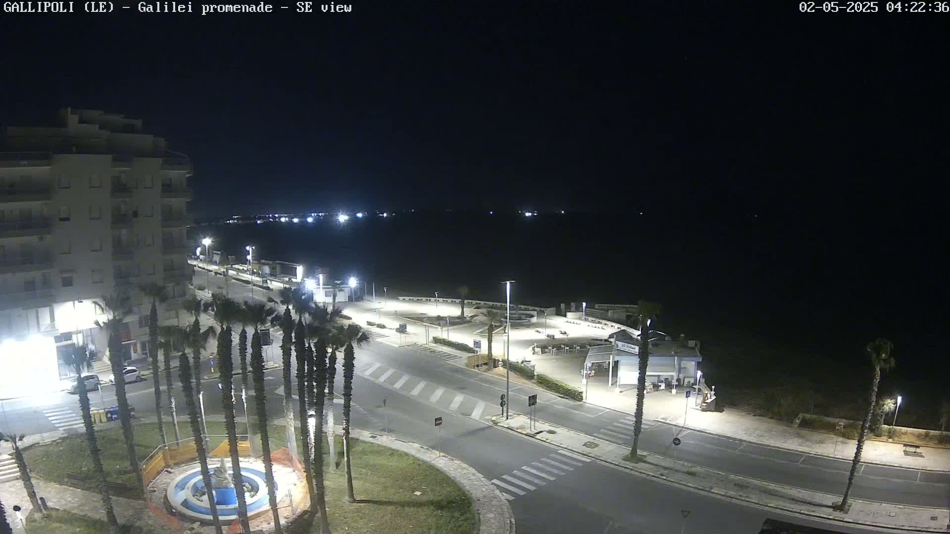 A nighttime, seaside view shows a mostly empty promenade with palm trees, a building, and a circular fountain, all under a clear, dark sky.