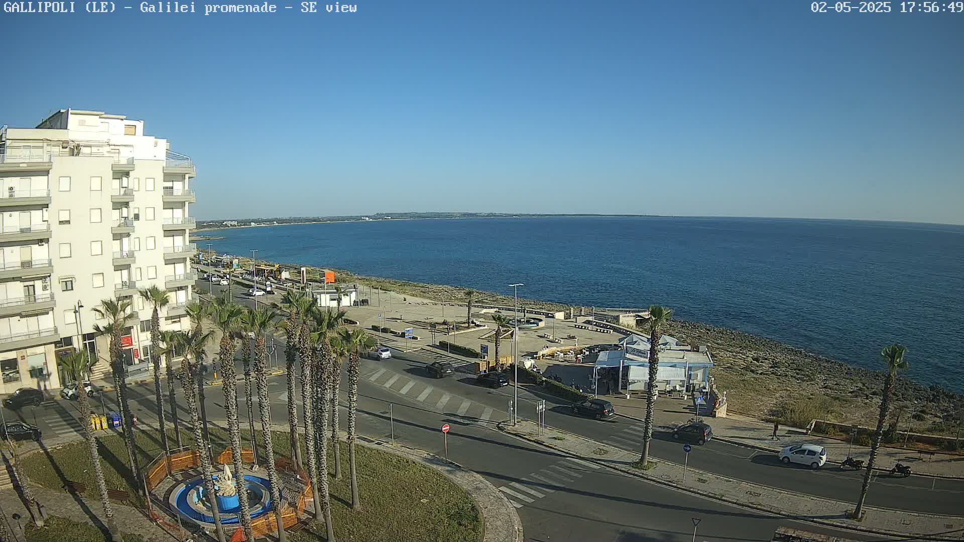 A seaside town scene on a sunny day shows a multi-story building, palm trees lining a road next to a promenade with several vehicles parked and driving, and a calm blue sea extending to a distant shoreline.