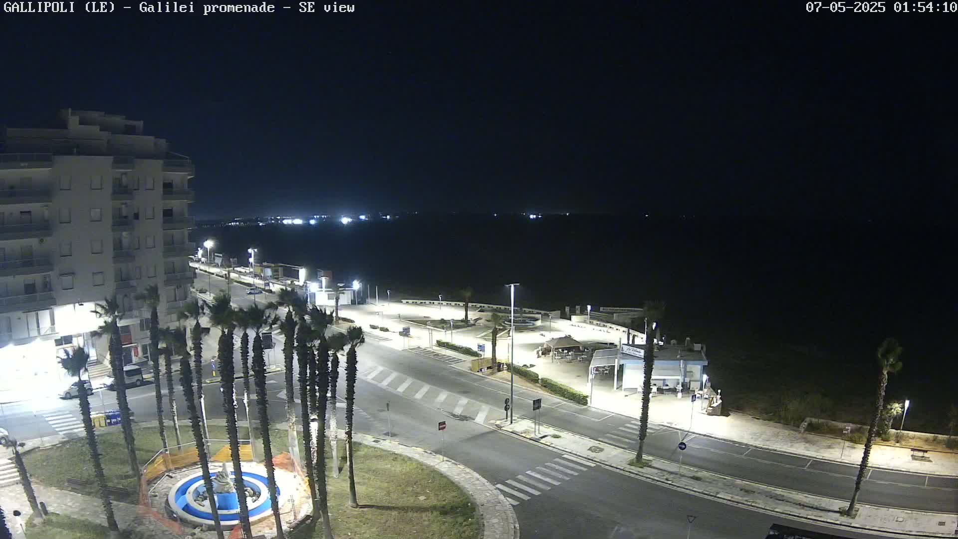 A nighttime, high-angle view of a seaside promenade shows a mostly empty road curving toward the dark ocean, lined with palm trees and a multi-story building.