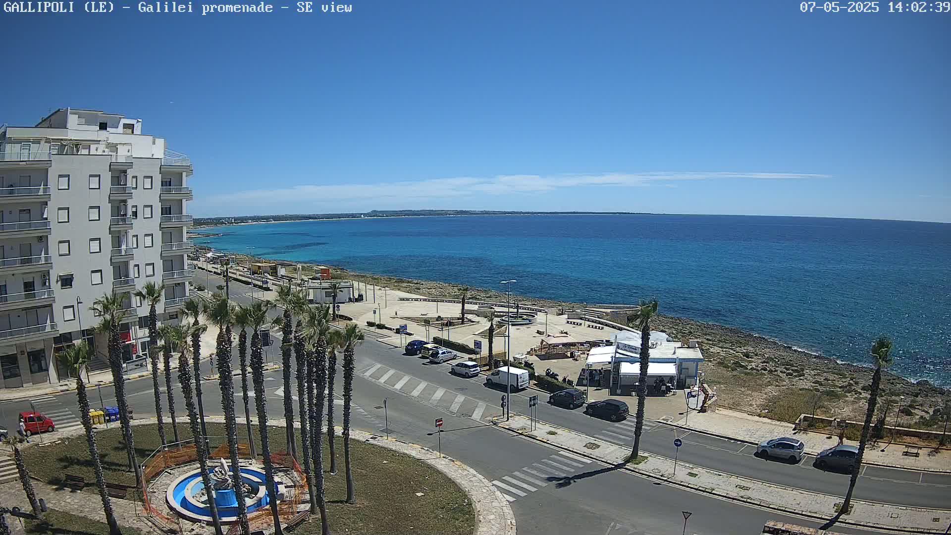 A seaside town on a sunny day, featuring a multi-story building, palm trees, a roadway, a small beach area with kiosks, and a calm, turquoise ocean in the background.