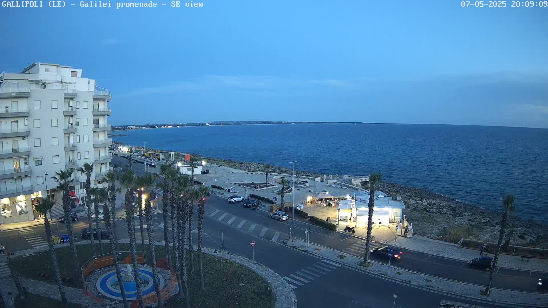 A seaside town at dusk shows a calm sea, a multi-story building, palm trees lining a street with cars, and a small beachfront kiosk.