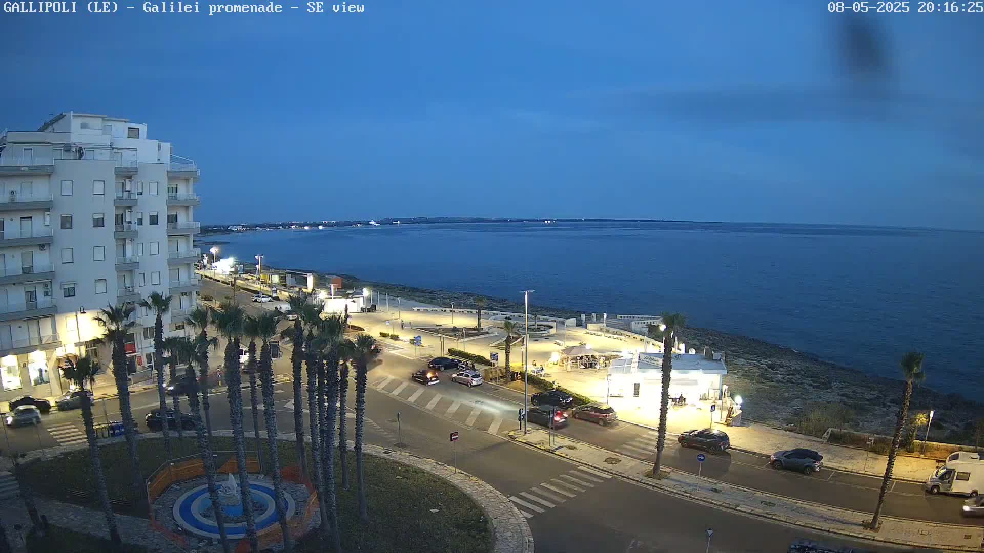 A seaside promenade at twilight, with palm trees lining a roadway next to a calm, dark-blue sea, and a multi-story building visible in the background.