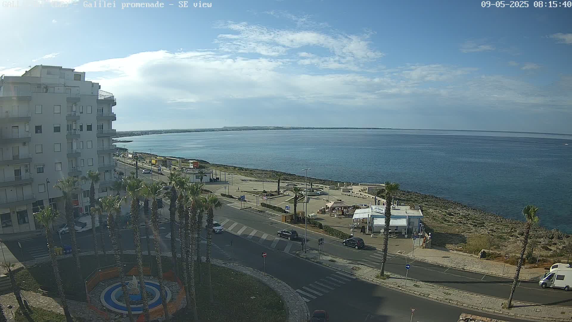A partly cloudy day reveals a seaside promenade with palm trees, a few cars, and buildings overlooking a calm sea.