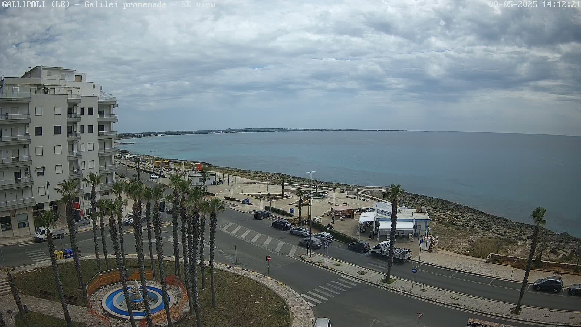 A partly cloudy day overlooks a coastal town with palm trees lining a street next to a calm sea.