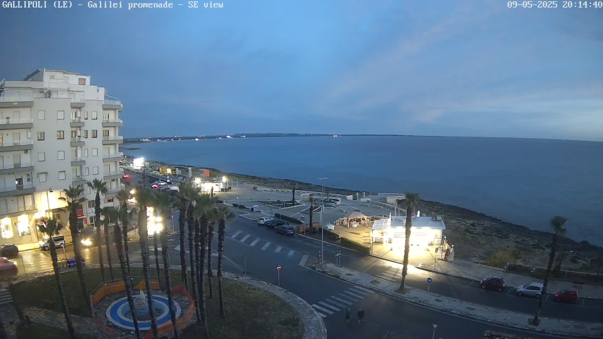 A coastal town at dusk, showing a calm sea under a mostly clear twilight sky, with palm trees lining a road near a multi-story building.
