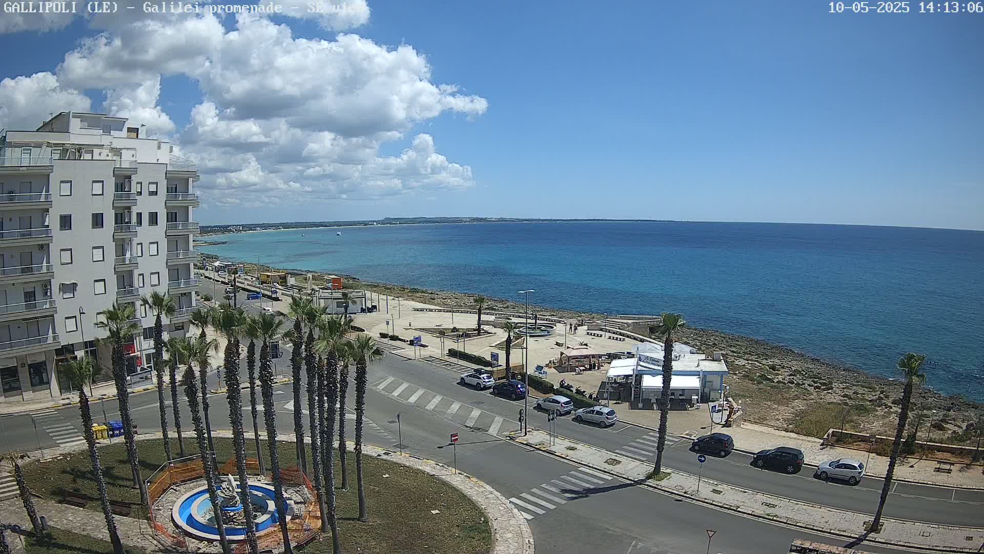 A partly cloudy day overlooks a coastal town with a calm, turquoise sea, palm trees lining a roadway near a building and a small beachfront plaza.
