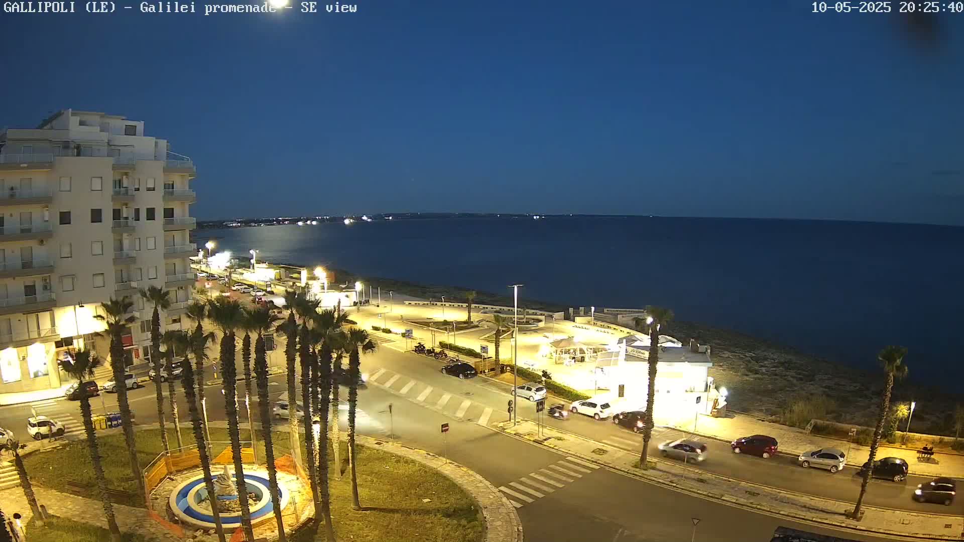 A nighttime seaside scene shows a calm, dark-blue sea, a town's illuminated streets with palm trees and parked cars, and a multi-story building near a circular plaza.