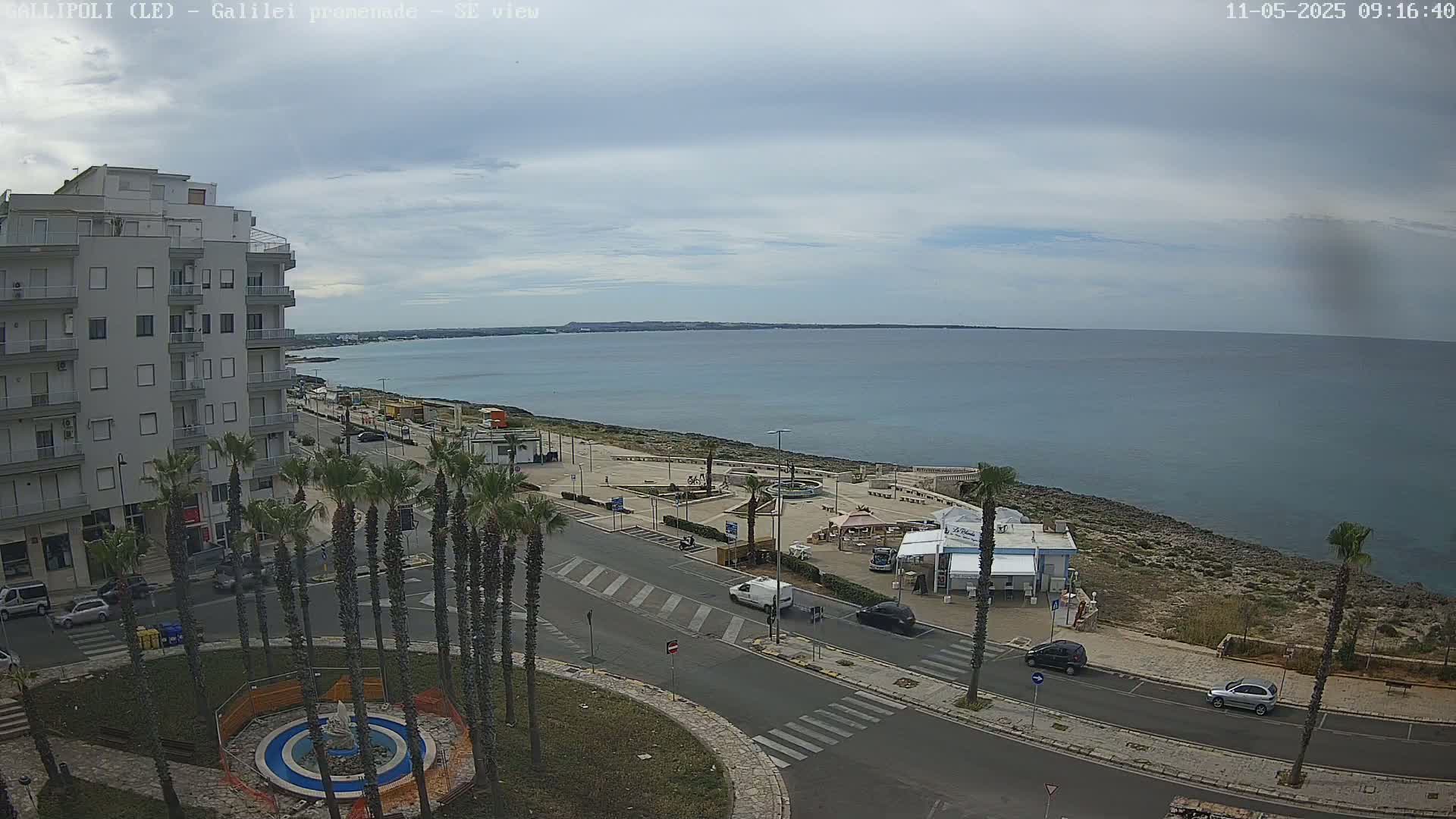 An overcast day reveals a seaside town with a multi-story building, palm trees lining a road leading to a waterfront promenade with a small kiosk, and a calm, blue sea in the background.
