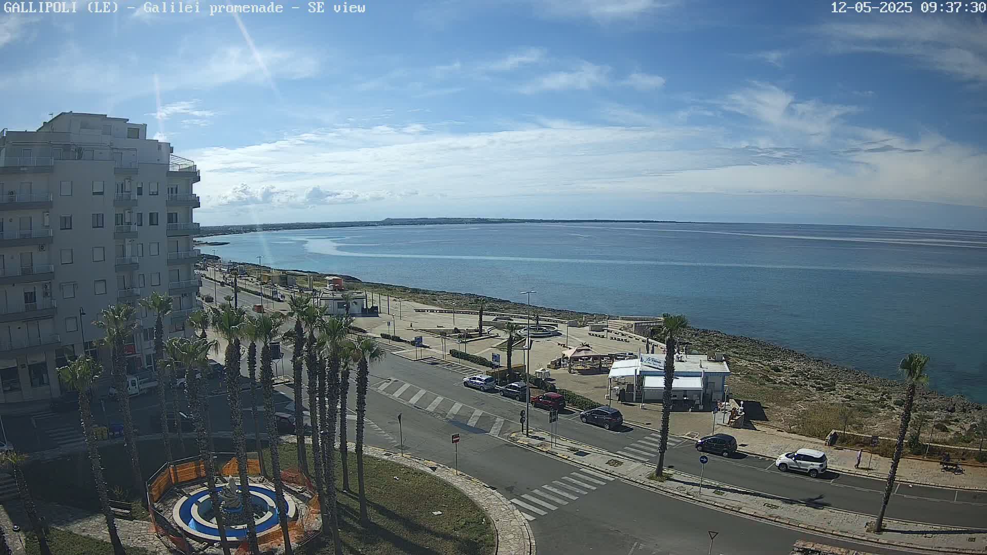 A seaside town scene under a partly cloudy sky shows a multi-story building, palm trees, a roadway, a beachfront plaza with a small fountain, and calm blue water extending to a distant shoreline.