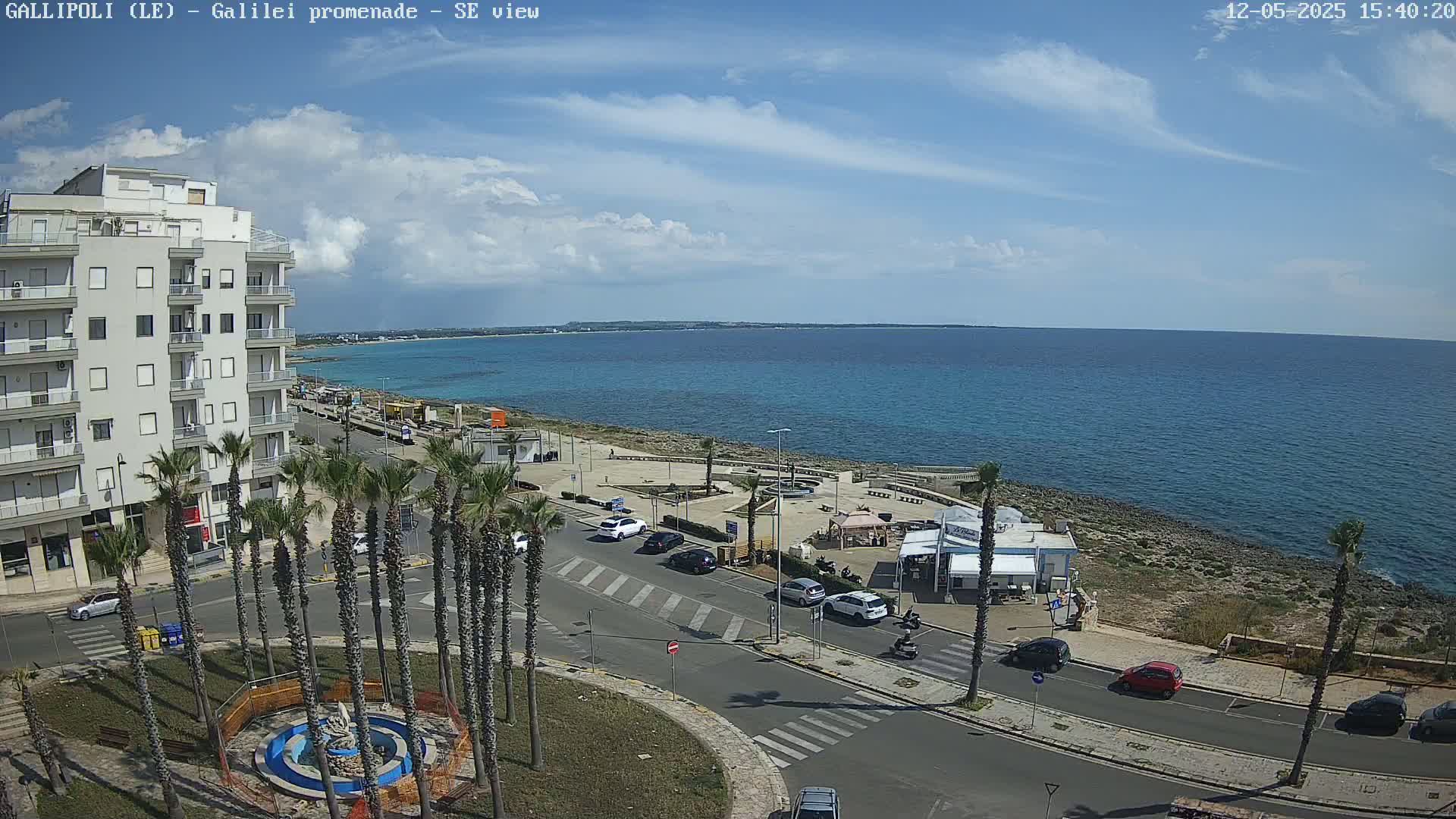 A partly cloudy sunny day overlooks a seaside town with palm trees lining a street next to a calm blue ocean.