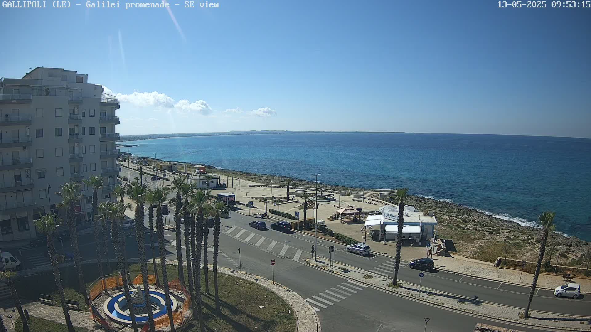 A seaside town on a sunny day with a few clouds, showing a coastal road with palm trees, buildings, and a calm blue sea.