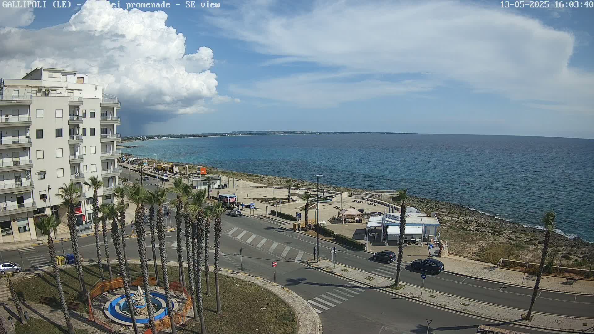 A coastal town scene on a partly cloudy day shows a multi-story building, a seaside promenade with palm trees, a roundabout, cars, and a calm blue ocean in the background.