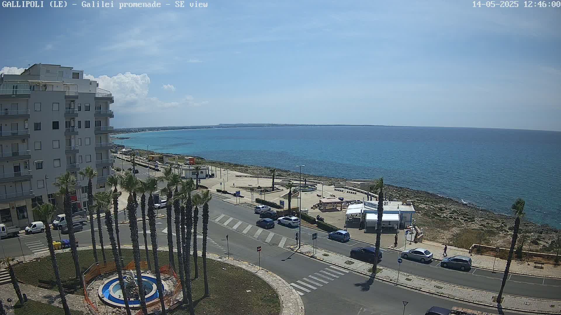 A seaside town scene on a partly cloudy day shows a multi-story building, palm trees lining a road leading to the ocean, cars parked along the street, and a small beachfront area with kiosks.