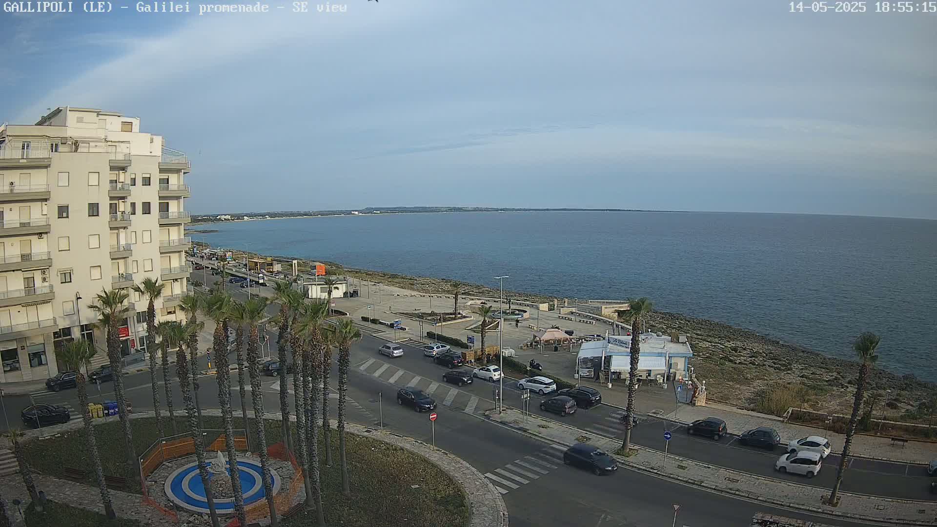 A seaside town on a partly cloudy day shows a calm ocean, a promenade with palm trees, cars on the road, and buildings.