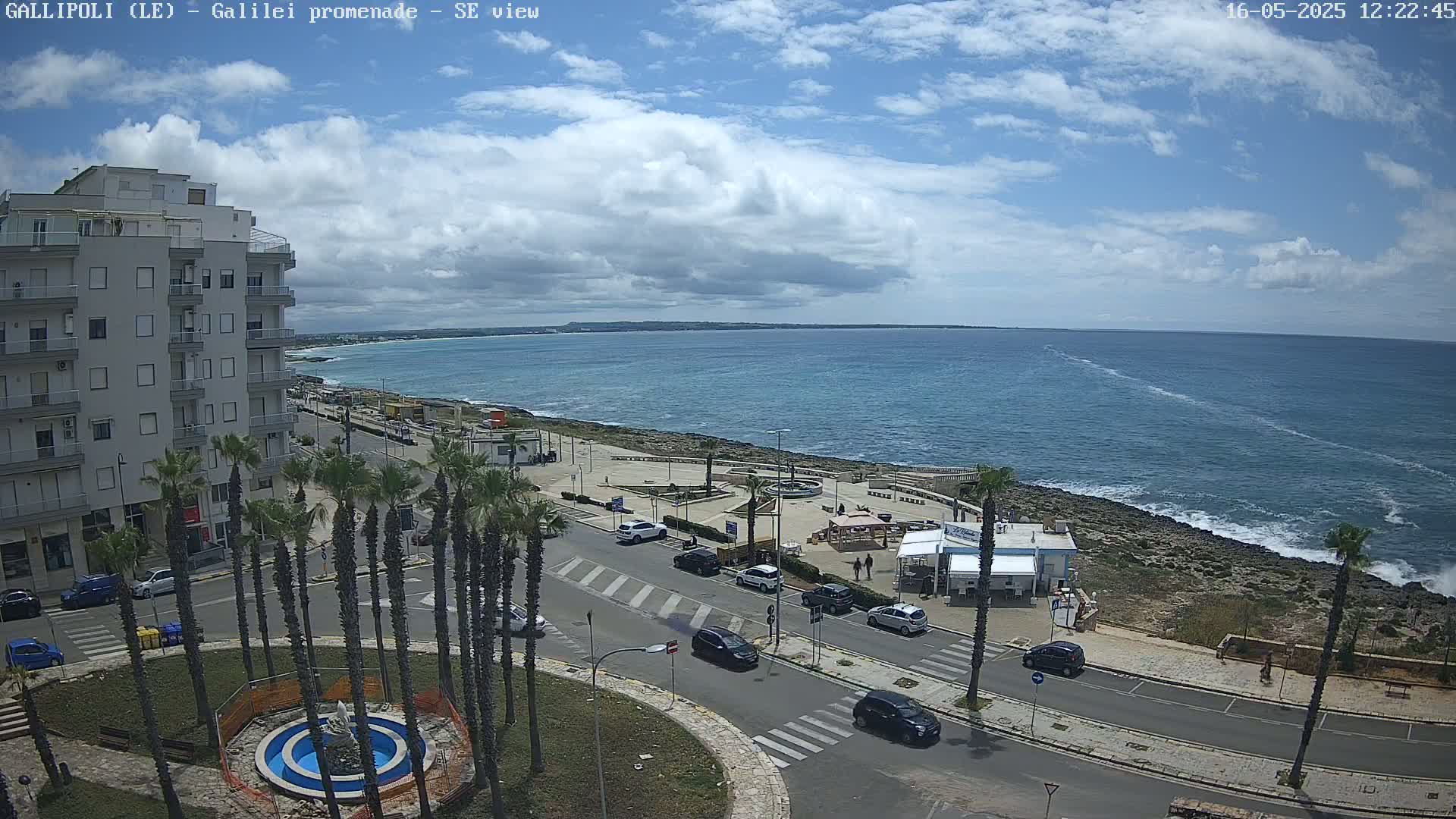 A partly cloudy day shows a seaside town with a building, palm trees, cars, and a promenade overlooking a calm ocean.