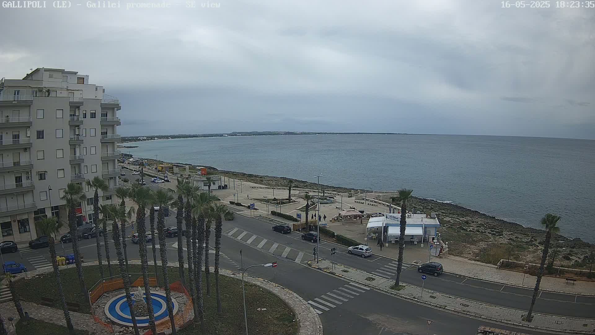 A seaside town under a cloudy sky features a multi-story building, palm trees lining a roadway, and a calm sea visible beyond a rocky shoreline.