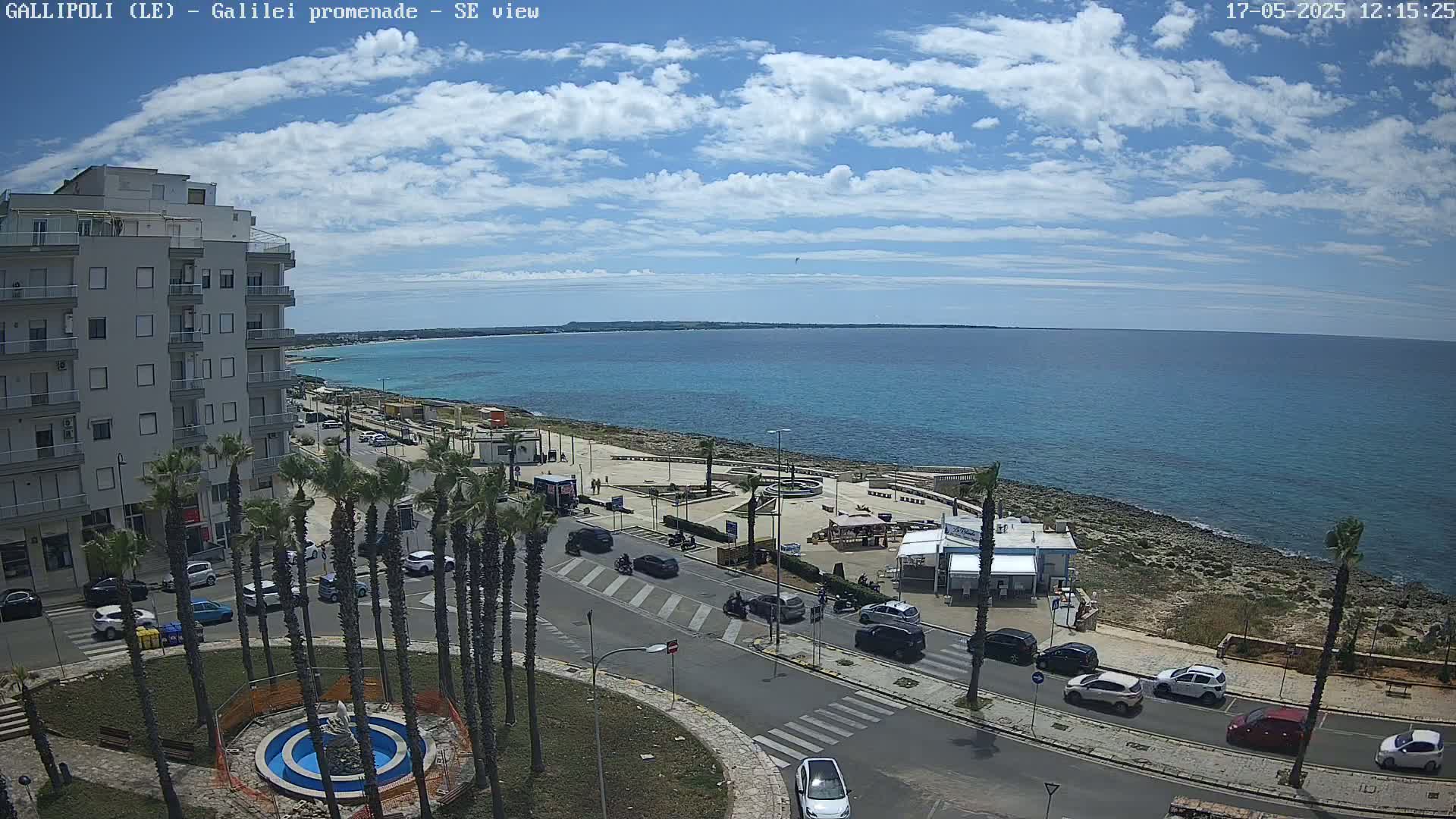 A partly sunny day reveals a seaside town with a promenade, palm trees, and cars parked along a road next to a turquoise ocean.