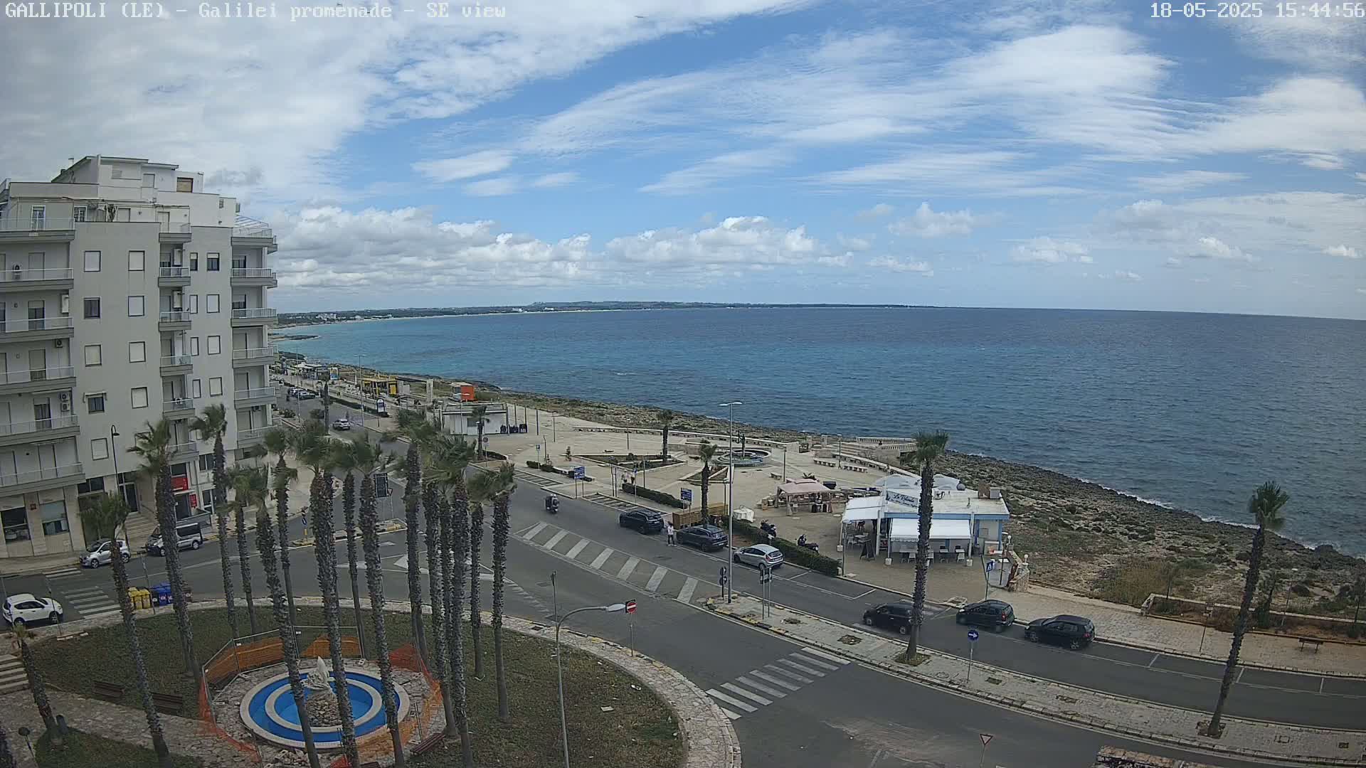 A coastal city scene under a partly cloudy sky shows a seaside promenade with palm trees, buildings, cars, and a small beach area next to a calm ocean.