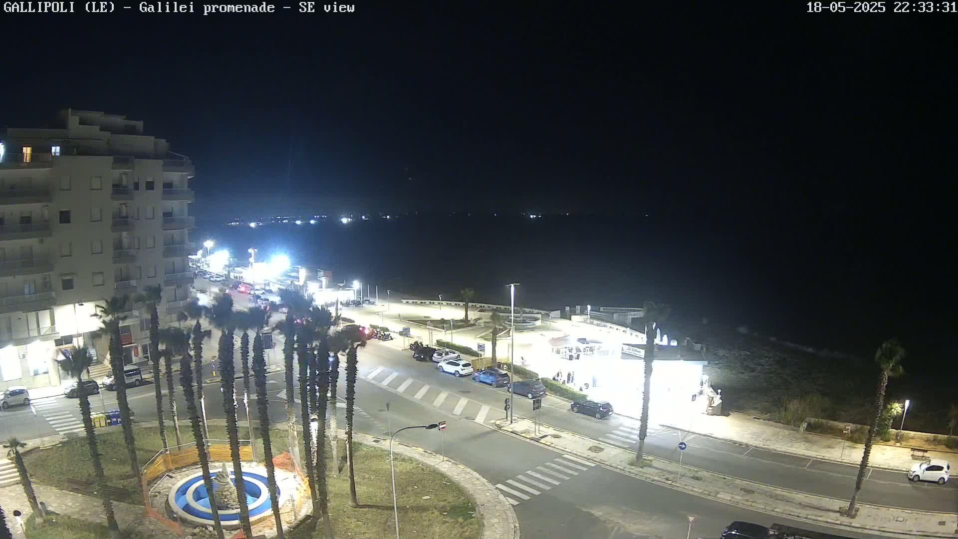 A nighttime, high-angle view of a coastal town with a multi-story building, palm trees, cars parked along roads, and a calm dark sea in the background.