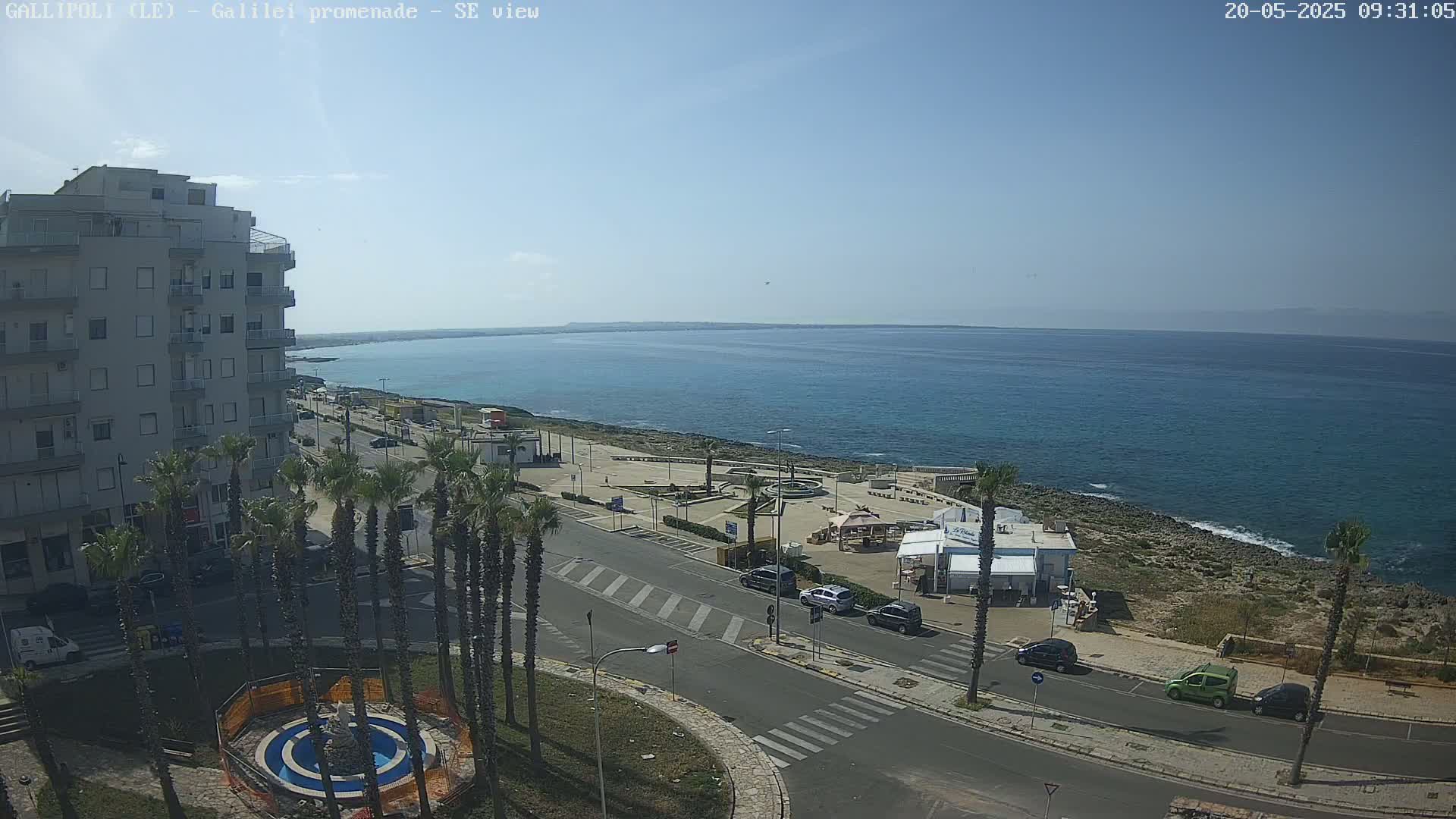 A seaside town scene on a sunny day shows a multi-story building, palm trees, a road, and a calm blue sea stretching to the horizon.