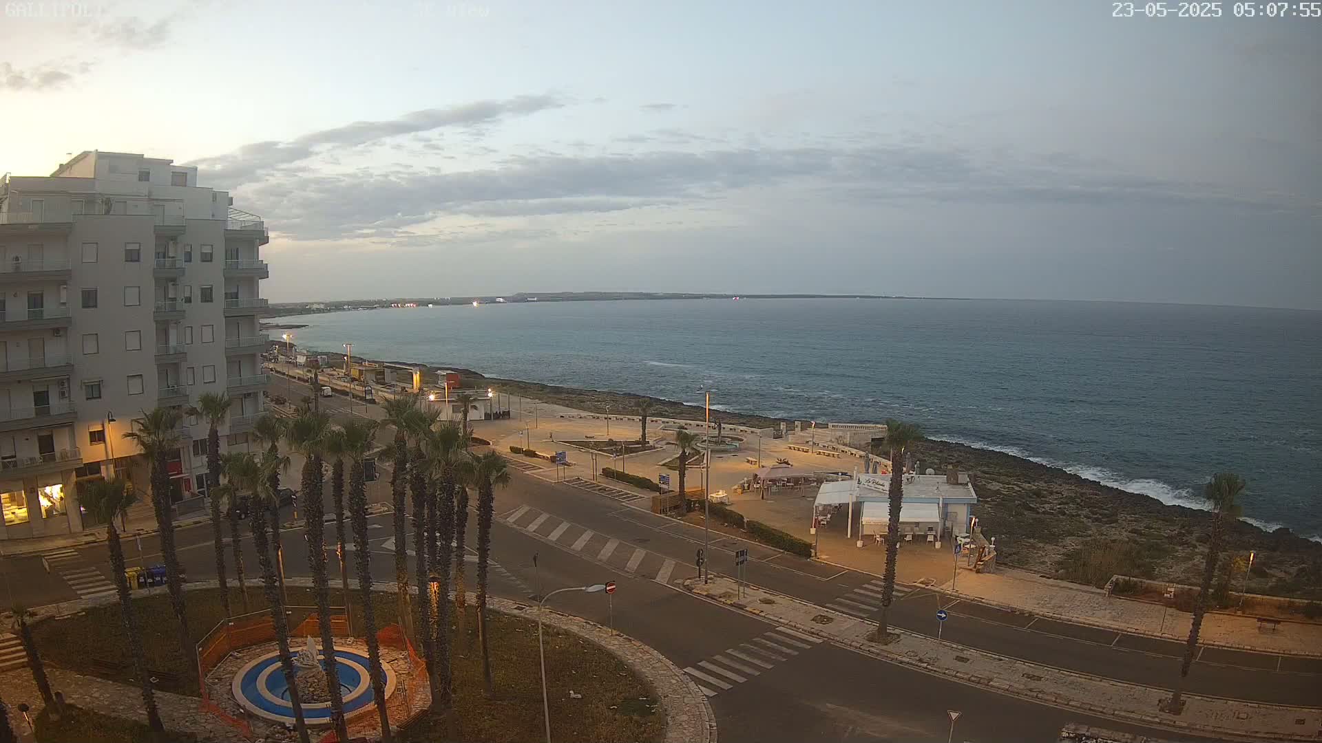 An overcast evening scene shows a coastal town with palm trees lining a road that leads to a rocky shoreline and calm sea.