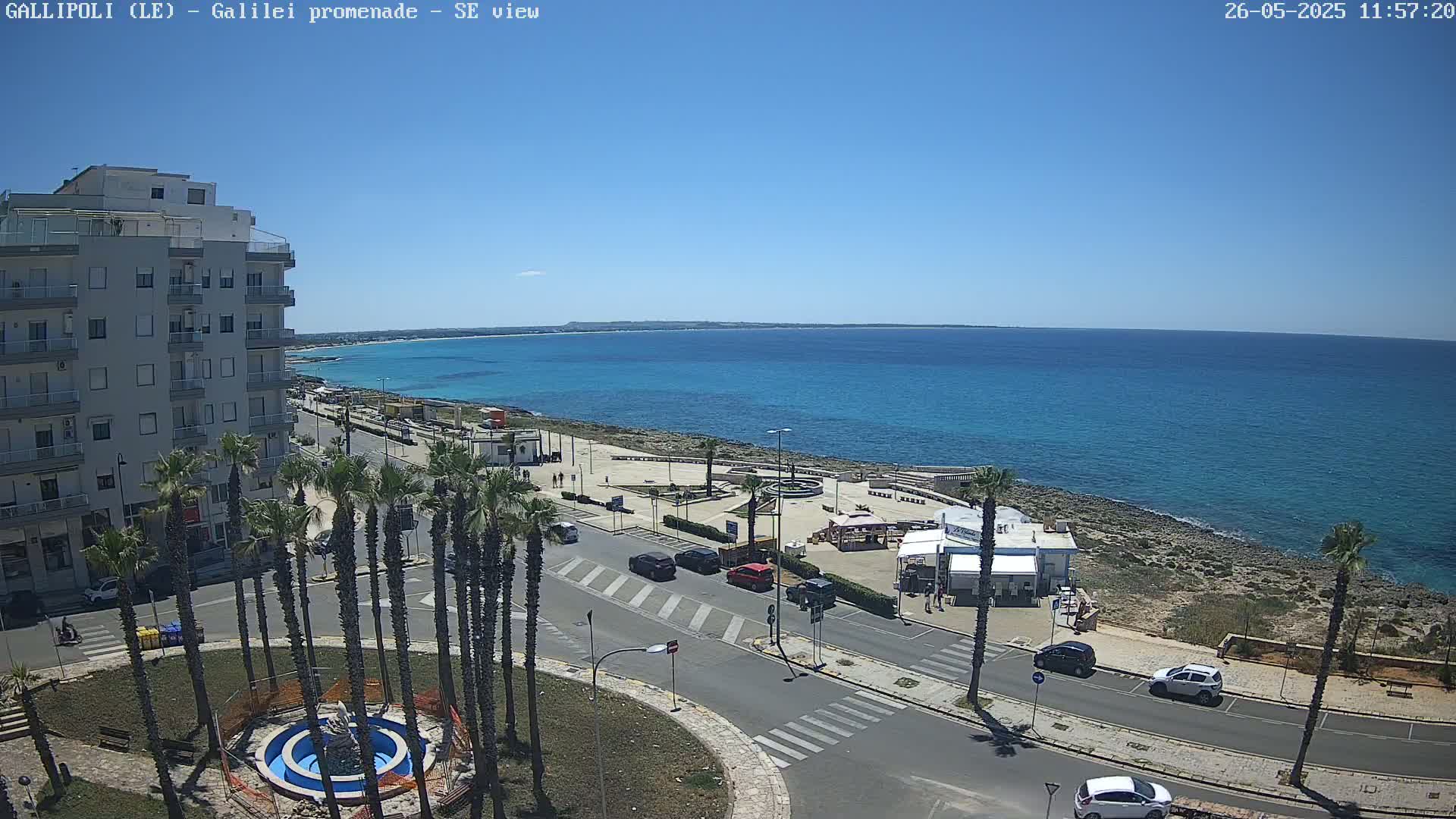 A seaside town on a sunny day features a multi-story building, palm trees lining a road near a waterfront plaza, and a calm turquoise sea extending to the horizon.