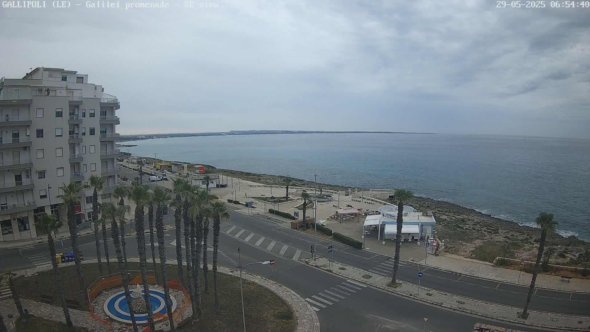 A mostly cloudy day reveals a seaside town with a multi-story building, palm trees lining a street leading to a waterfront plaza, and a calm sea extending to a distant shoreline.