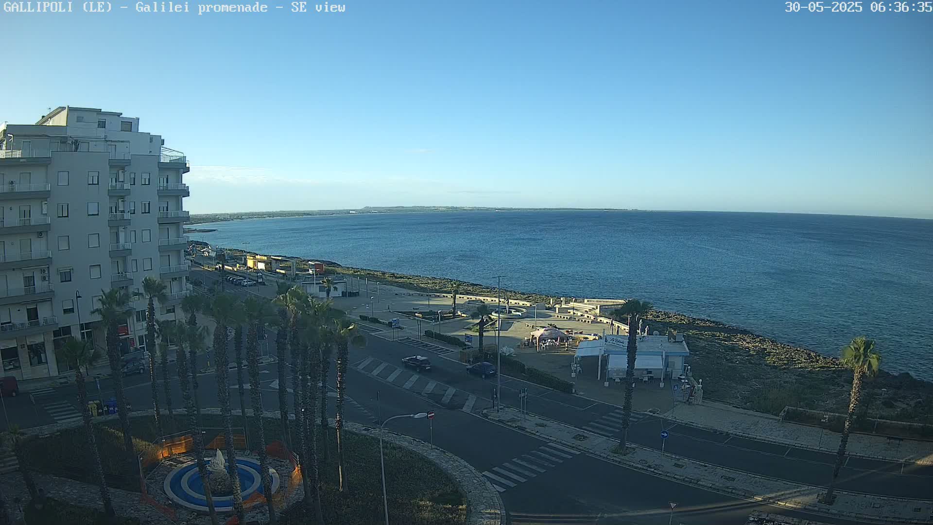 A seaside town under a clear blue sky shows a paved promenade next to a calm ocean, palm trees lining a street near a multi-story building.