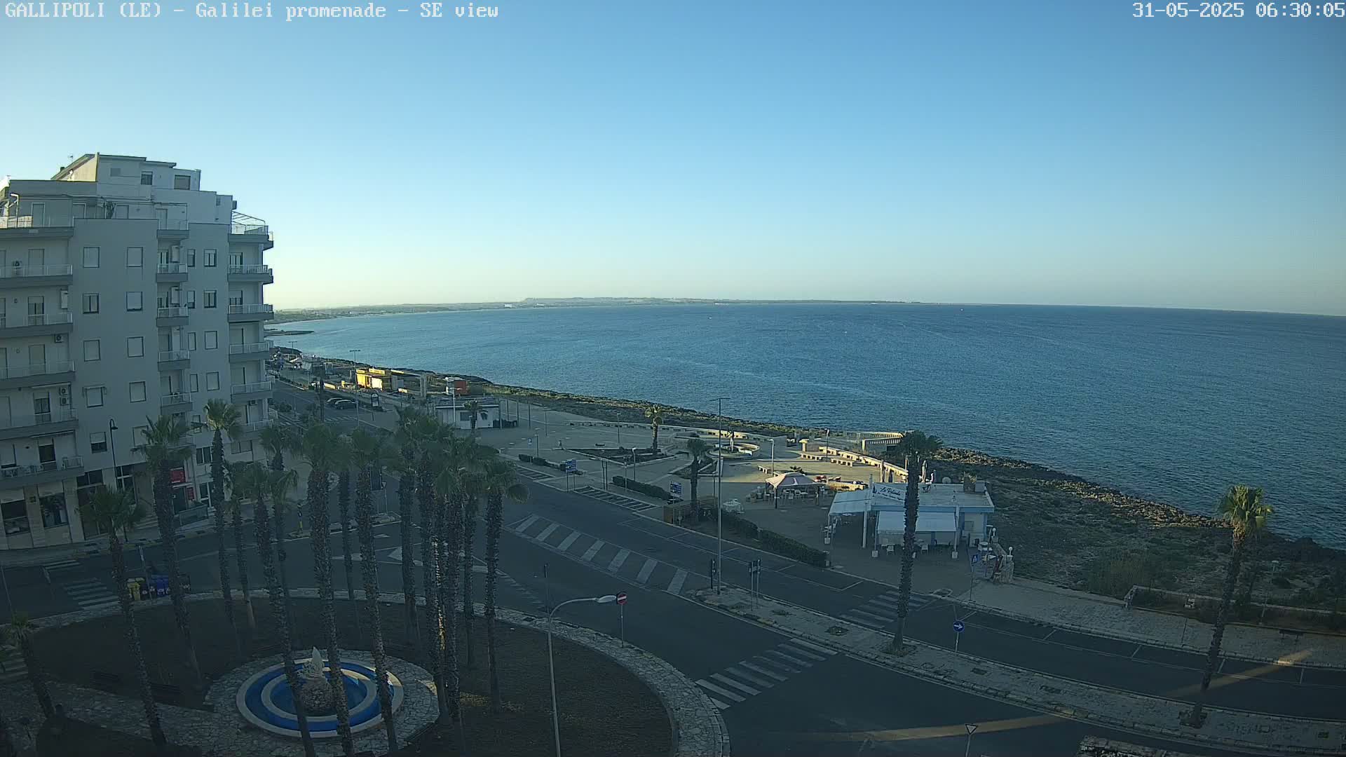 A seaside town scene on a clear day shows a calm blue ocean, a paved promenade with palm trees, and a multi-story building.