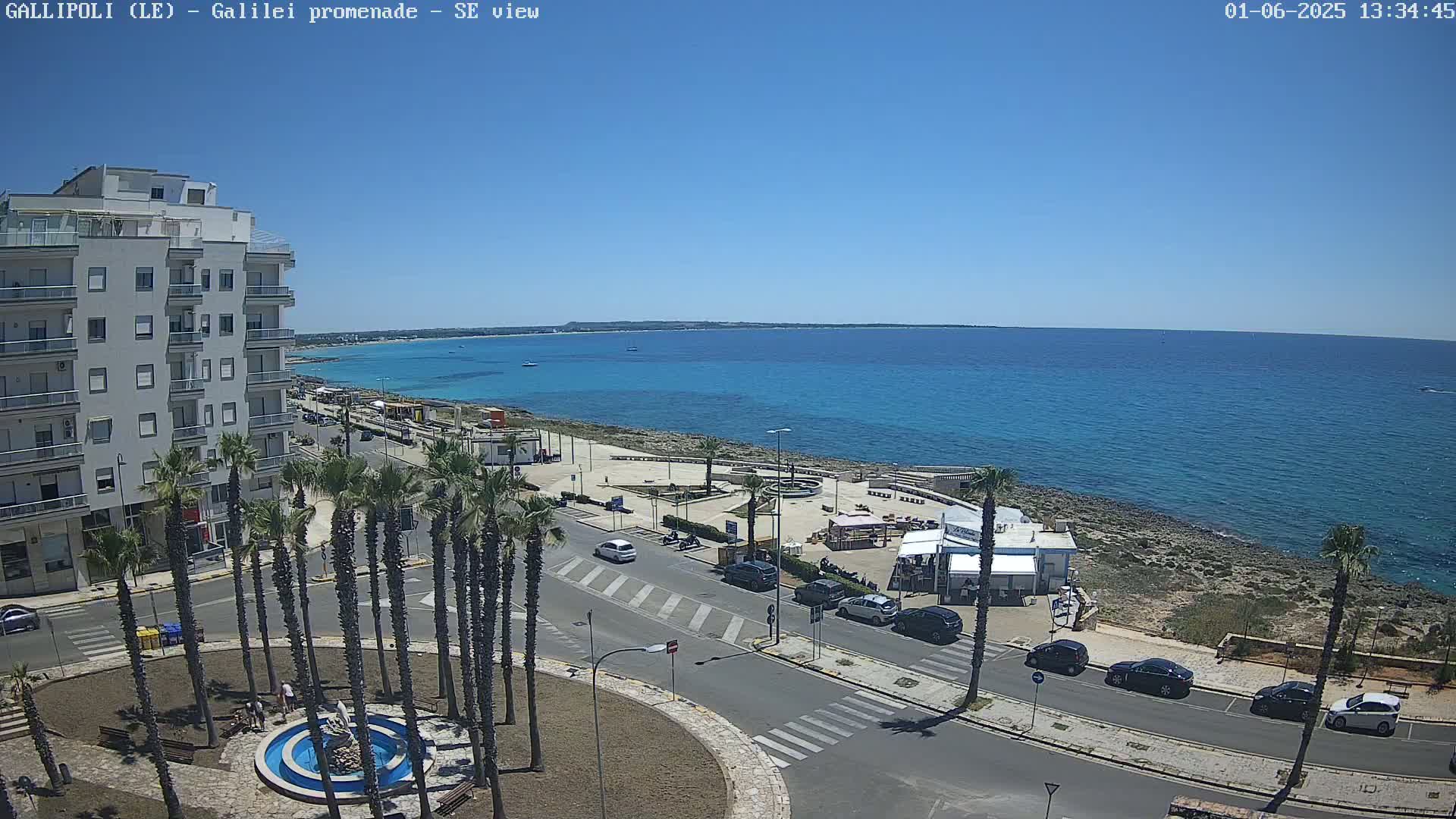 A seaside town on a sunny day, featuring a multi-story building, palm trees, a road with cars, and a rocky shoreline with turquoise water extending to the horizon.