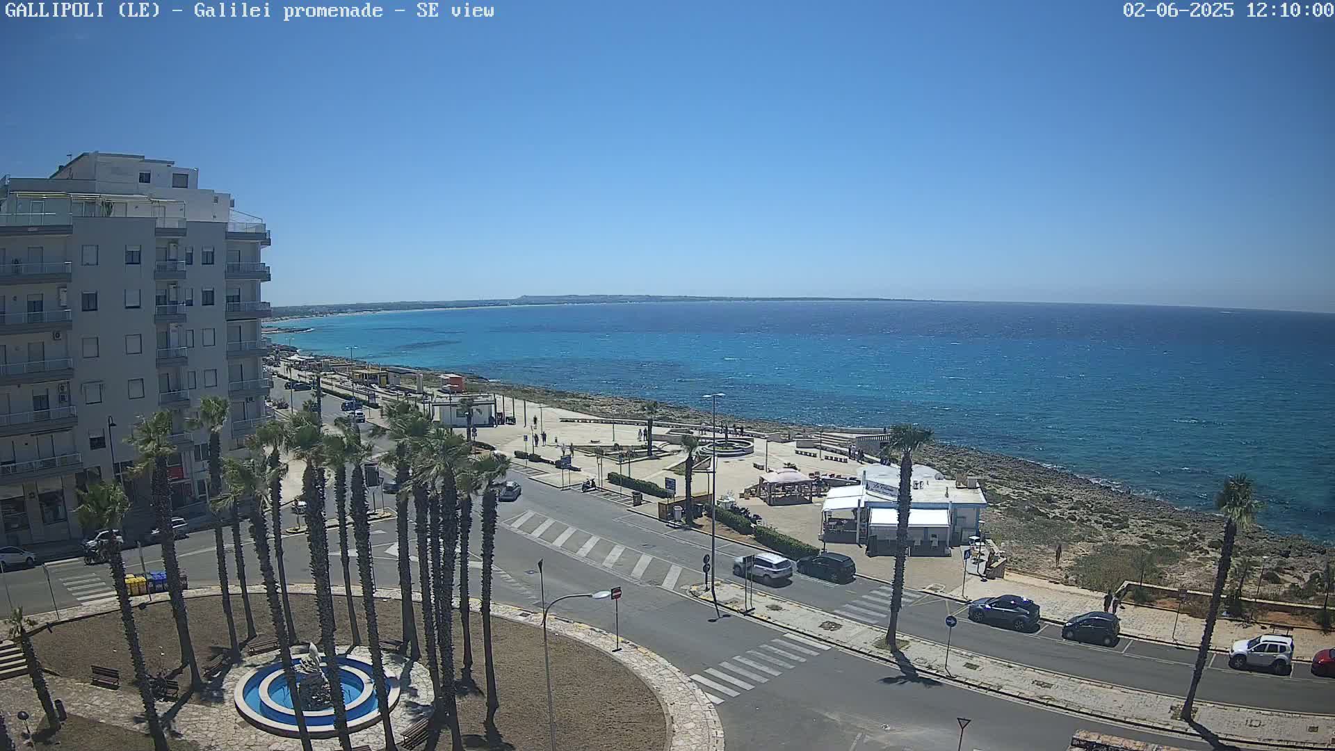 A seaside promenade scene on a sunny day shows a multi-story building, palm trees, a circular fountain, parked cars, and a calm, blue ocean.