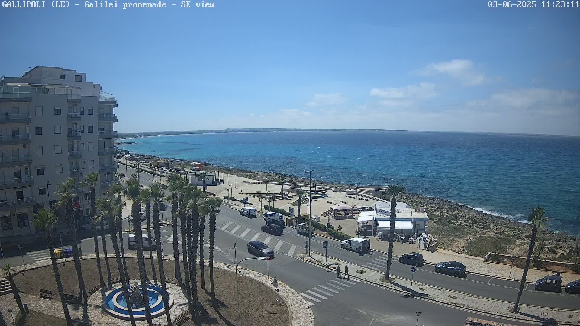 A seaside town scene on a sunny day shows a road lined with palm trees leading to a waterfront promenade with turquoise water and a multi-story building in the background.