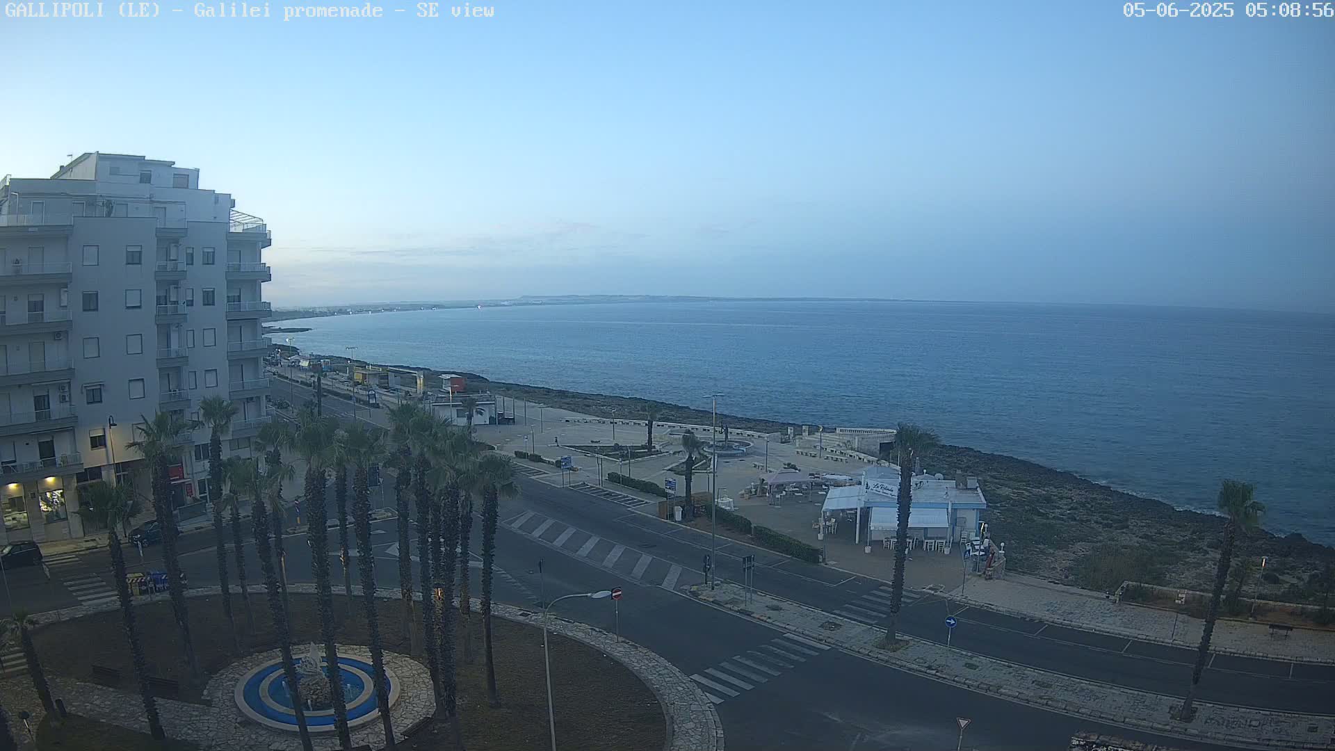A calm, clear morning reveals a coastal cityscape with palm trees lining a roadway that overlooks a tranquil sea.