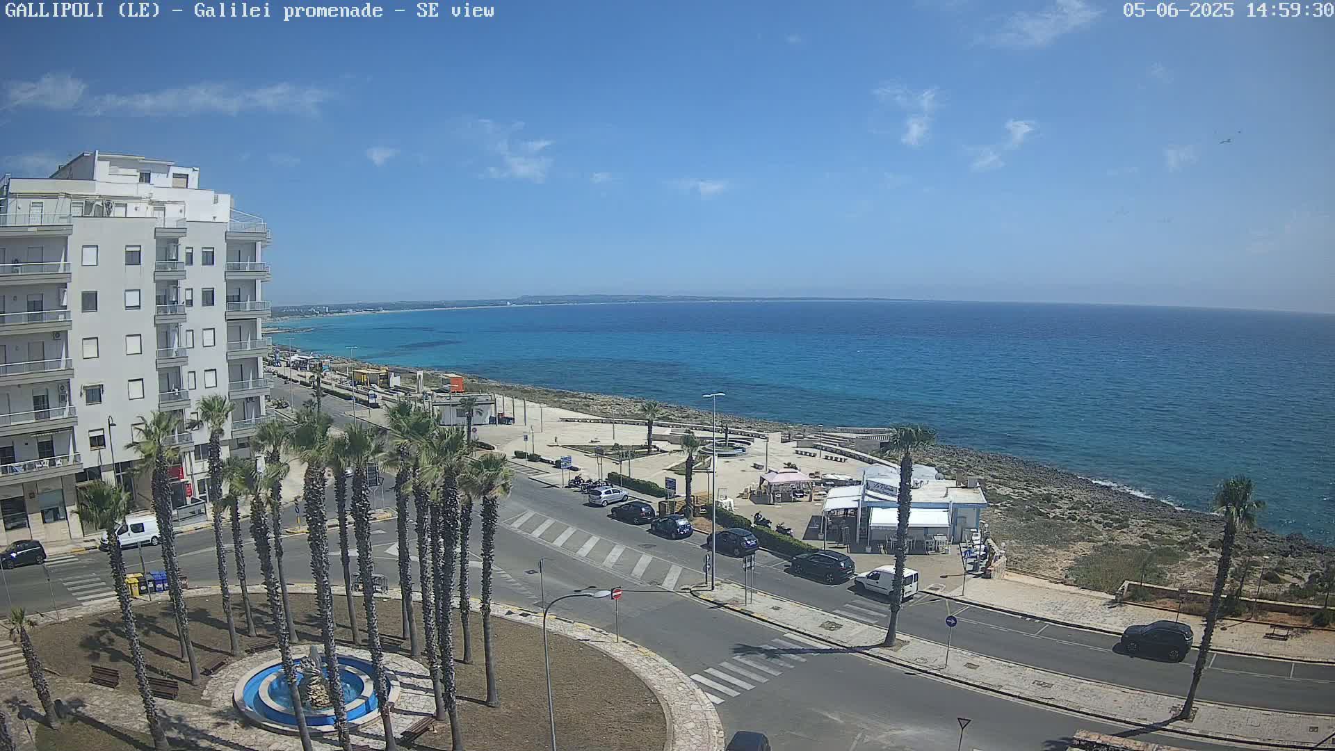 A seaside town scene on a sunny day shows a multi-story building, palm trees lining a street leading to a promenade with a few parked cars and small beachfront businesses, overlooking a calm, blue ocean.