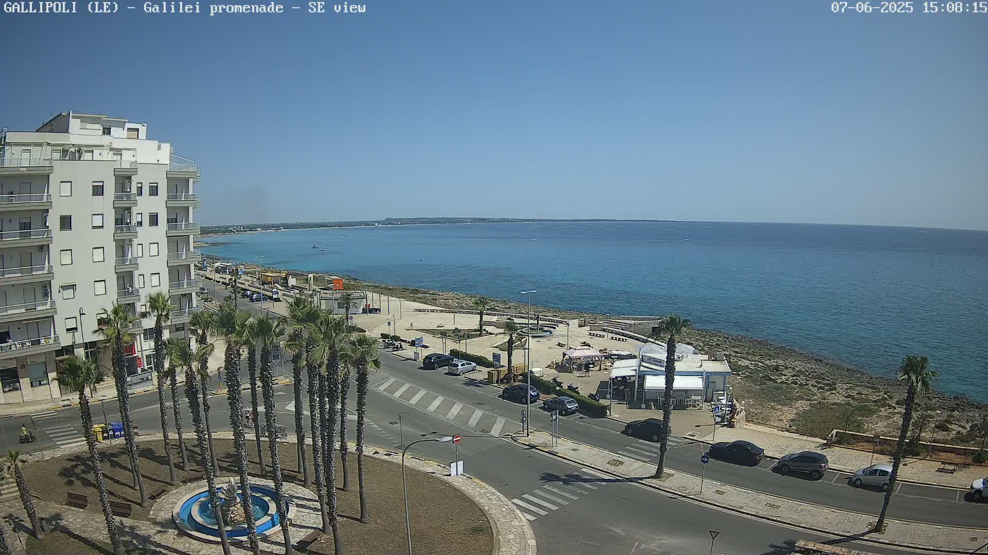 A clear, sunny day overlooking a coastal town with a calm blue sea, palm trees lining a street next to a building, and cars parked along the roadside near beachfront cafes.