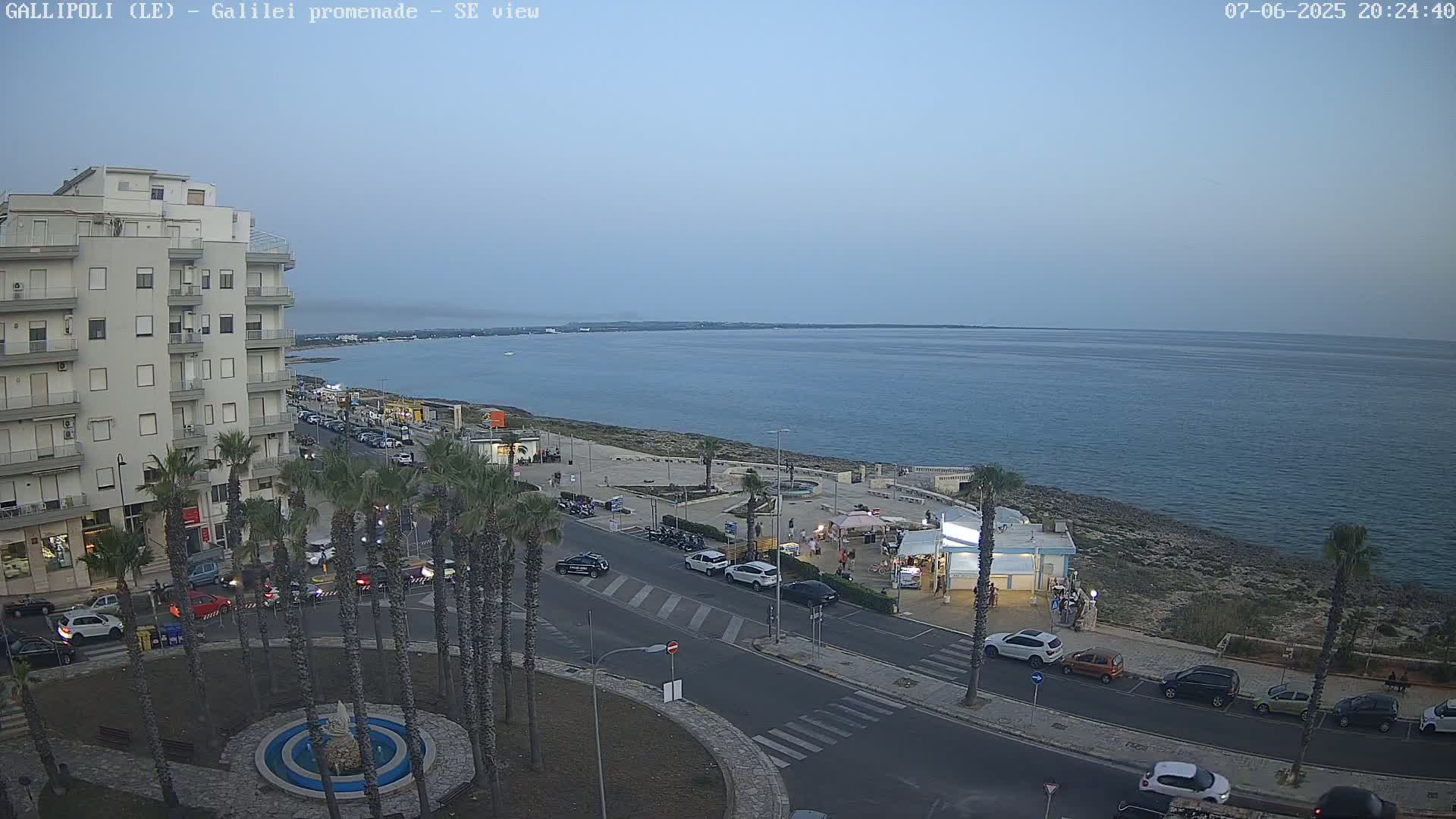 A seaside town at dusk, showing a calm ocean, a promenade with palm trees, parked cars, and several buildings, under a clear, pale sky.