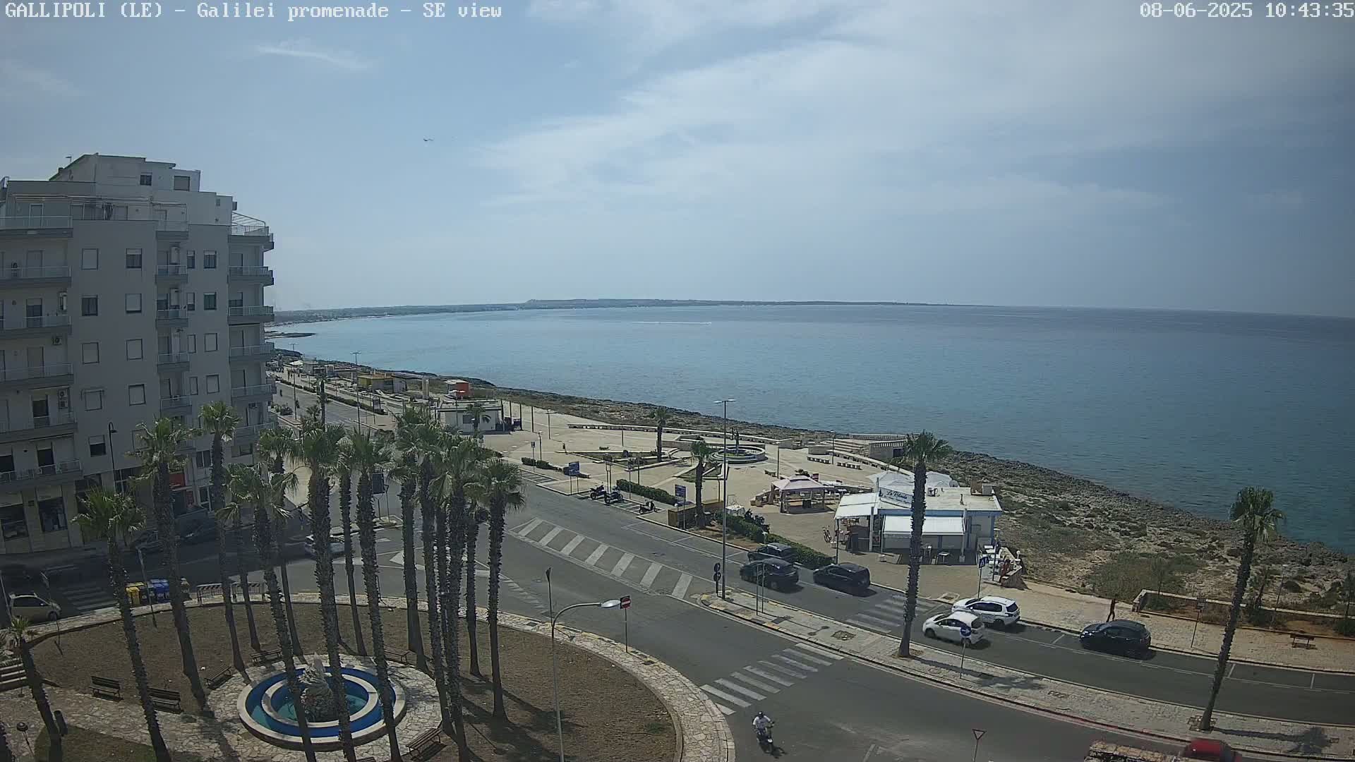 A partly cloudy day reveals a seaside town with a promenade, palm trees, and a circular fountain; cars are parked along the road and a few are driving.